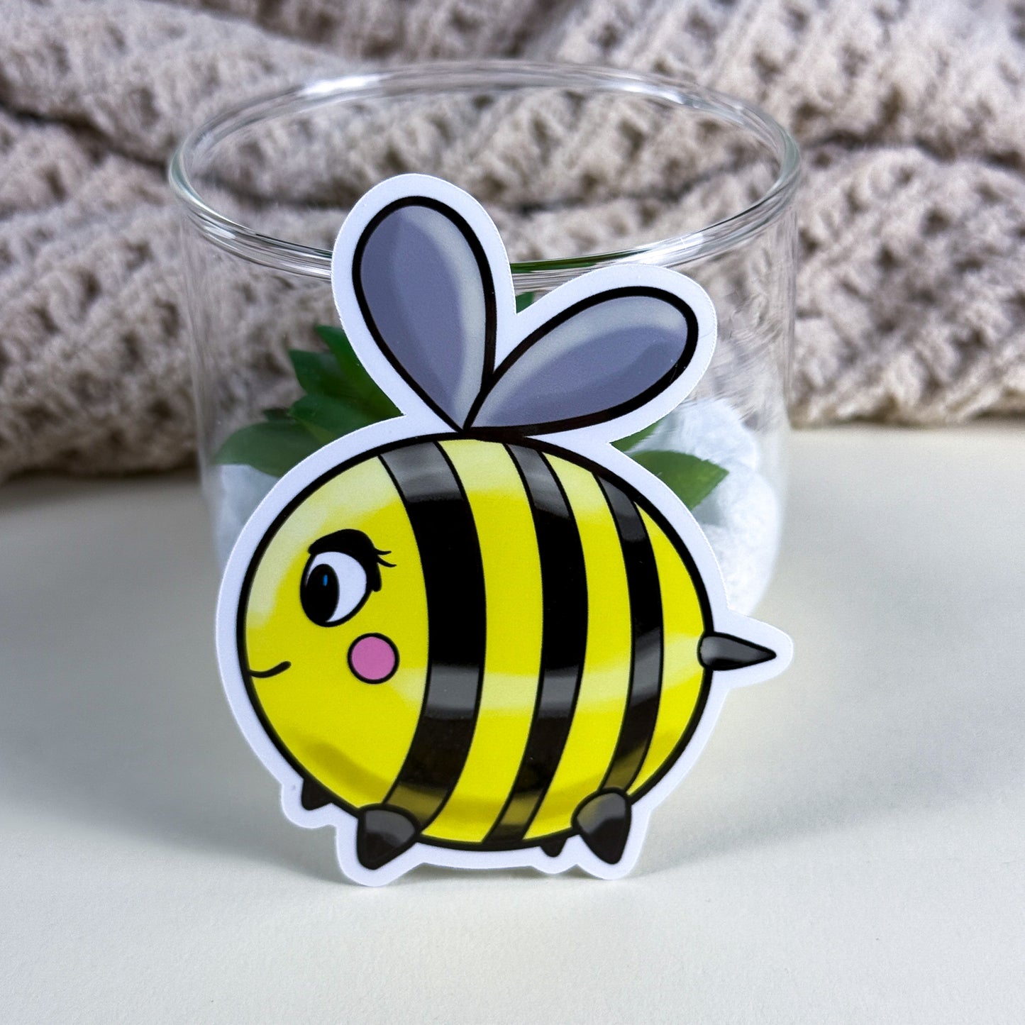 • Cute hand-drawn bee vinyl sticker in holographic finish on a white background, Kittlins • Close-up of waterproof Kittlins bee sticker applied to a teal water bottle • Bee sticker in standard glossy vinyl next to a journal and pen on a desk