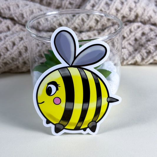 • Cute hand-drawn bee vinyl sticker in holographic finish on a white background, Kittlins • Close-up of waterproof Kittlins bee sticker applied to a teal water bottle • Bee sticker in standard glossy vinyl next to a journal and pen on a desk
